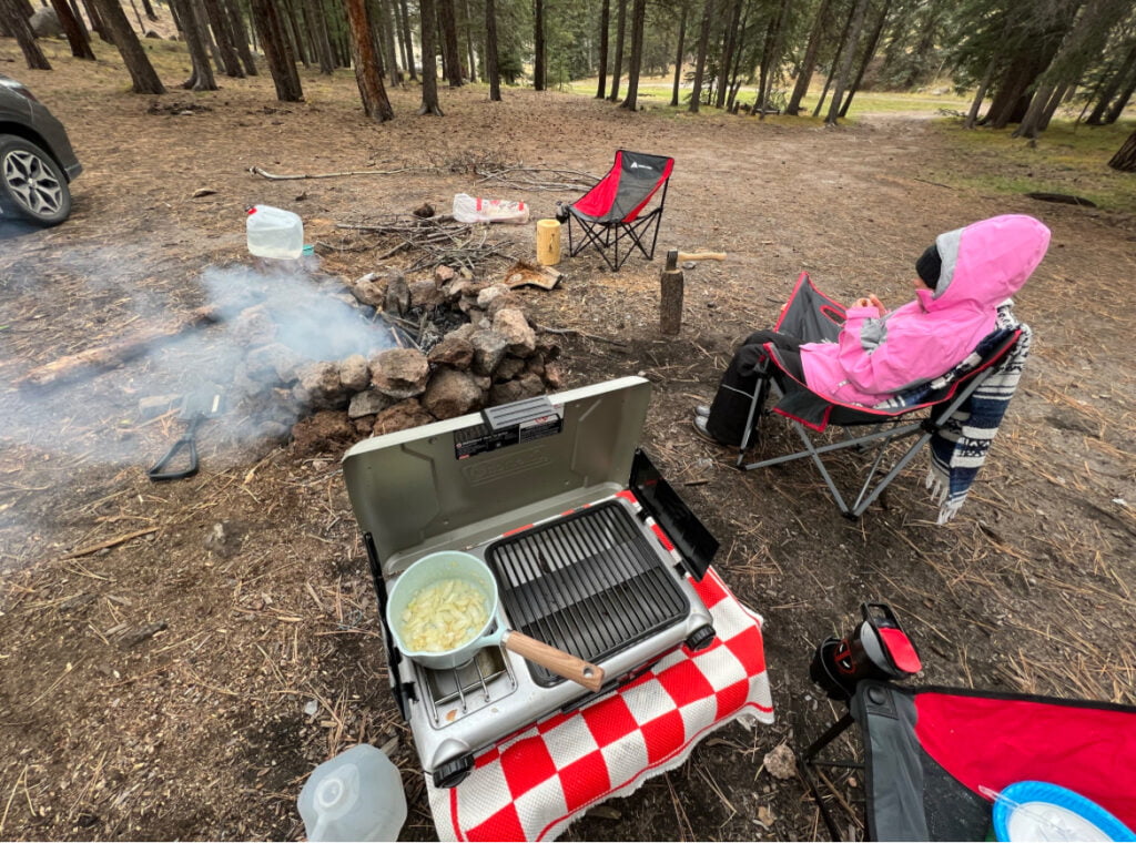 Most forgotten camping items that should definitely be on your checklist! 4 A girl in a pink jacket sits next to a campfire in the woods.