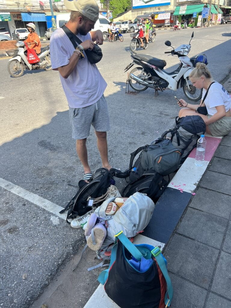 A man and a woman sitting on the side of the road in Thailand with their luggage.