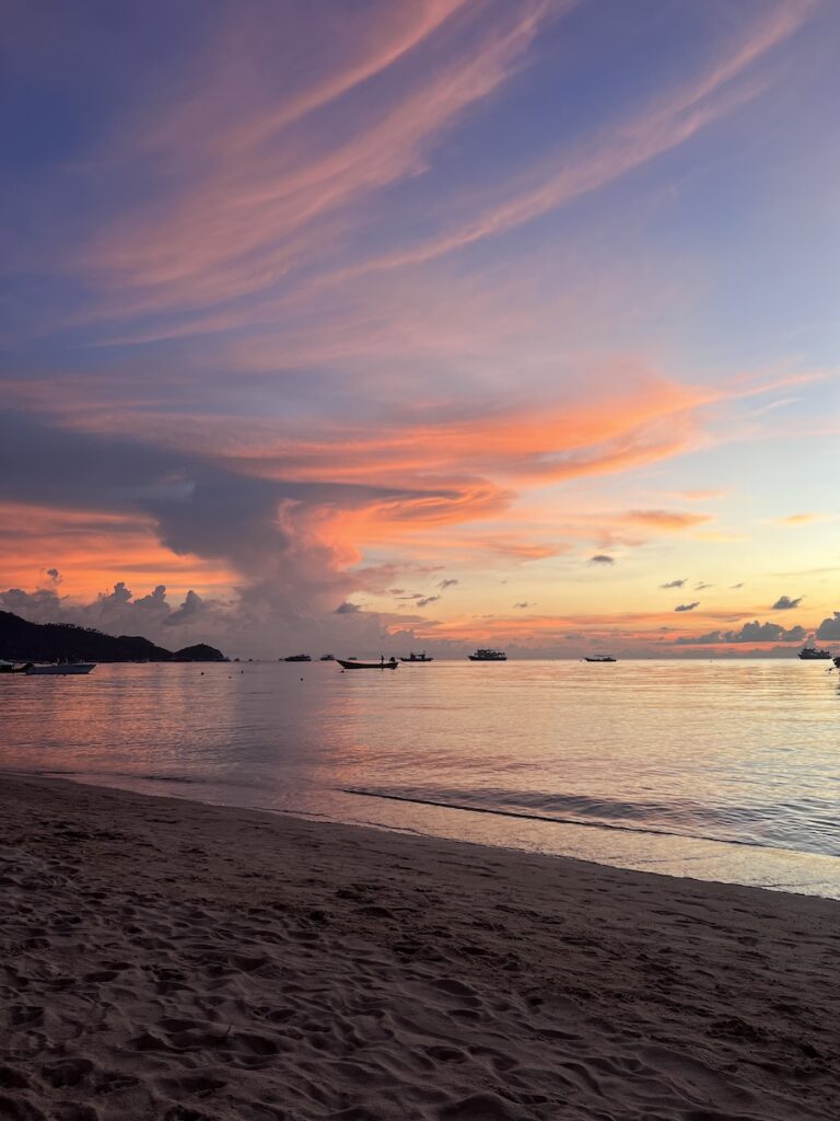 A sunset on a beach in Thailand with boats in the water.